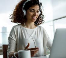 A woman of color with curly hair smiles, wears headphones and interacts with her laptop computer