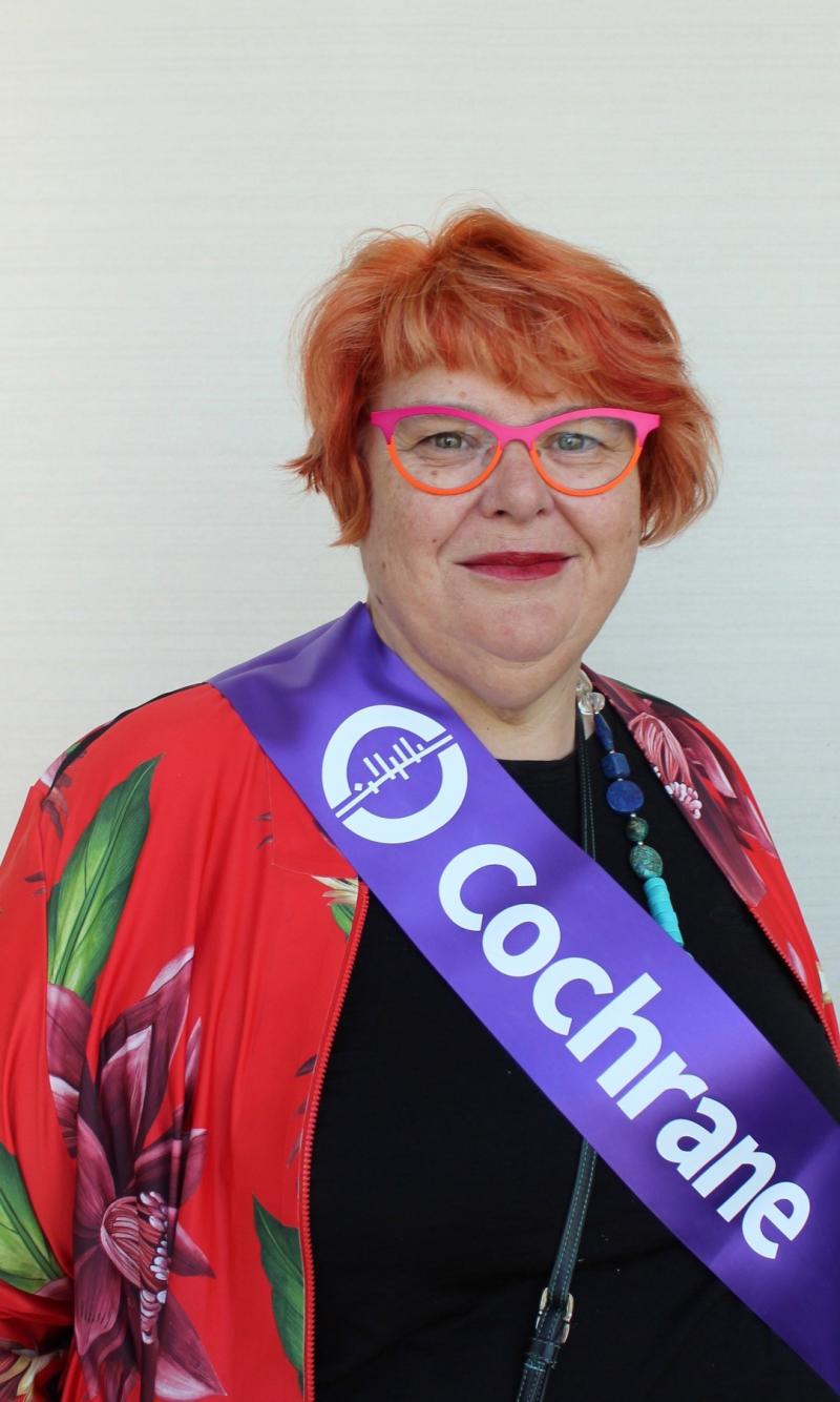 Catherine Marshall, a white woman with short red hair and pink and orange glasses, stands against a wall wearing a red, flowered jacket and a purple Cochrane sash