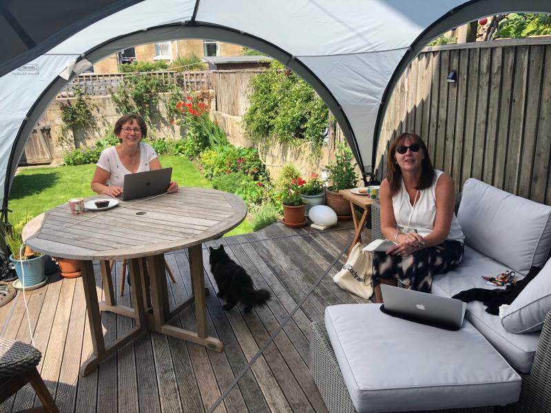 Two smiling women sit in a garden at a patio table