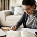 A Black woman with short curly hair sits at a table and writes in a notebook