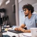a Black man with short curly hair and a beard sits at a computer