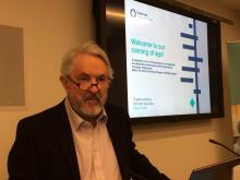 Richard Wormald, a white man with white hair and a white goatee, looks at the camera while standing in front of a presentation screen and wearing glasses