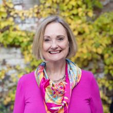 Helen Worthington, a white woman with chin-length blonde hair, smiles at the camera in a bright pink blouse and floral scarf