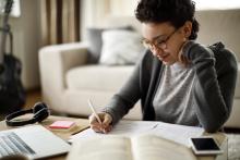 A Black woman with short curly hair sits at a table and writes in a notebook