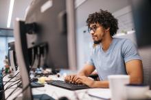 a Black man with short curly hair and a beard sits at a computer