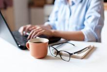 A woman sits at a laptop and is typing with a cup of coffee, notebook, and eyeglasses beside her