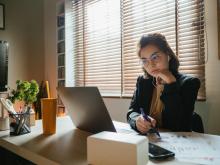 A woman looks at her computer with headphones in while she takes notes