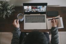 person sits at a computer desk with a pad of paper, coffee, and laptop which has a grid of meeting participants on it