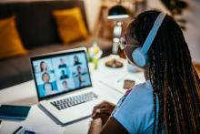 a black woman sits at a desk with her laptop open before her, with a grid of people's faces on her screen